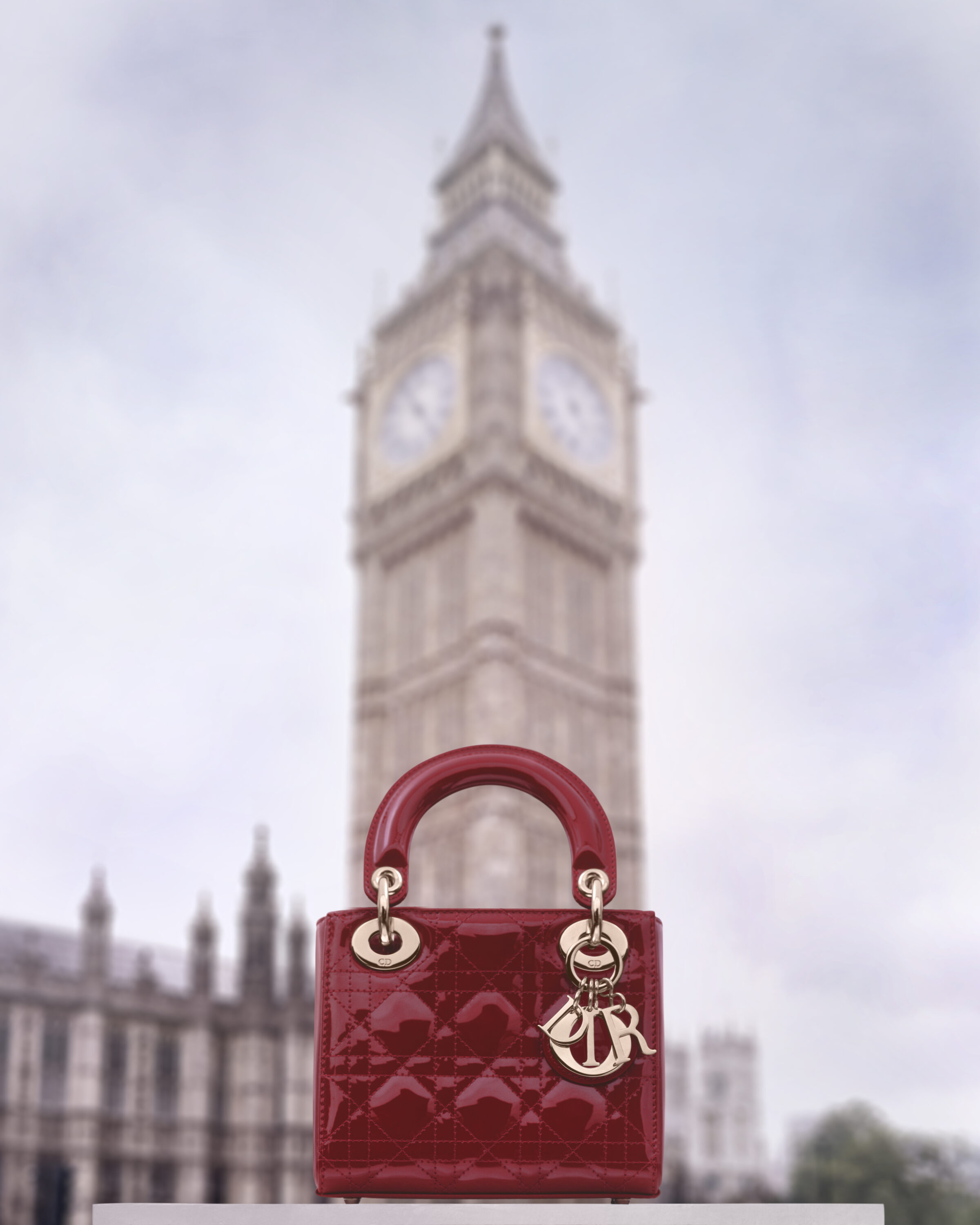 Red patent leather Lady Dior handbag positioned in front of a blurred London clock tower under an overcast sky.