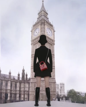 Model standing with back to camera wearing black dress and tall boots carrying a red Dior handbag in front of Big Ben.