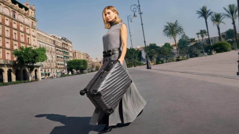 Woman in grey top holding a hardside suitcase walking on an urban plaza in Mexico City for Tumi Spring 2025 campaign