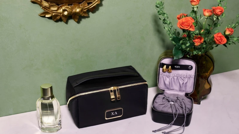 Black cosmetic pouch with gold zippers and monogram plaque next to an open jewelry case and perfume bottle