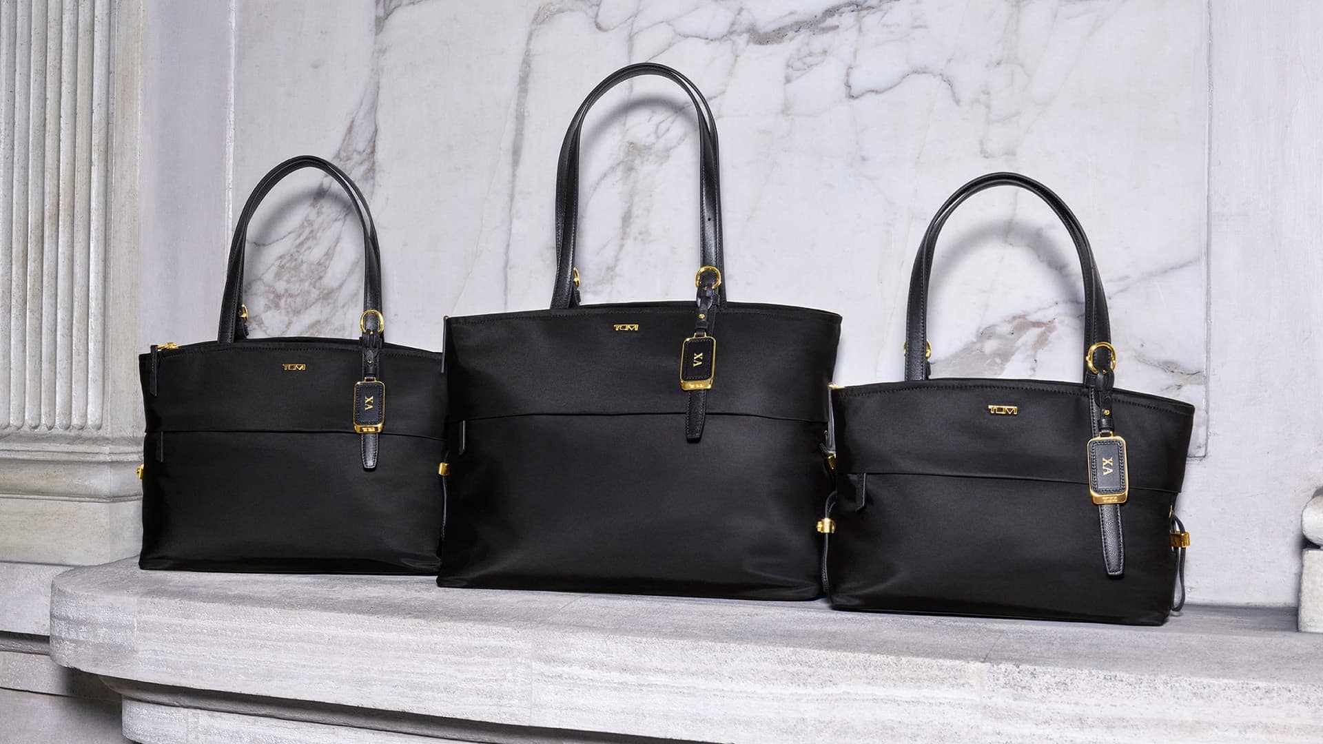 Three black Tumi tote bags displayed on a light marble stone surface against an architectural marble background.