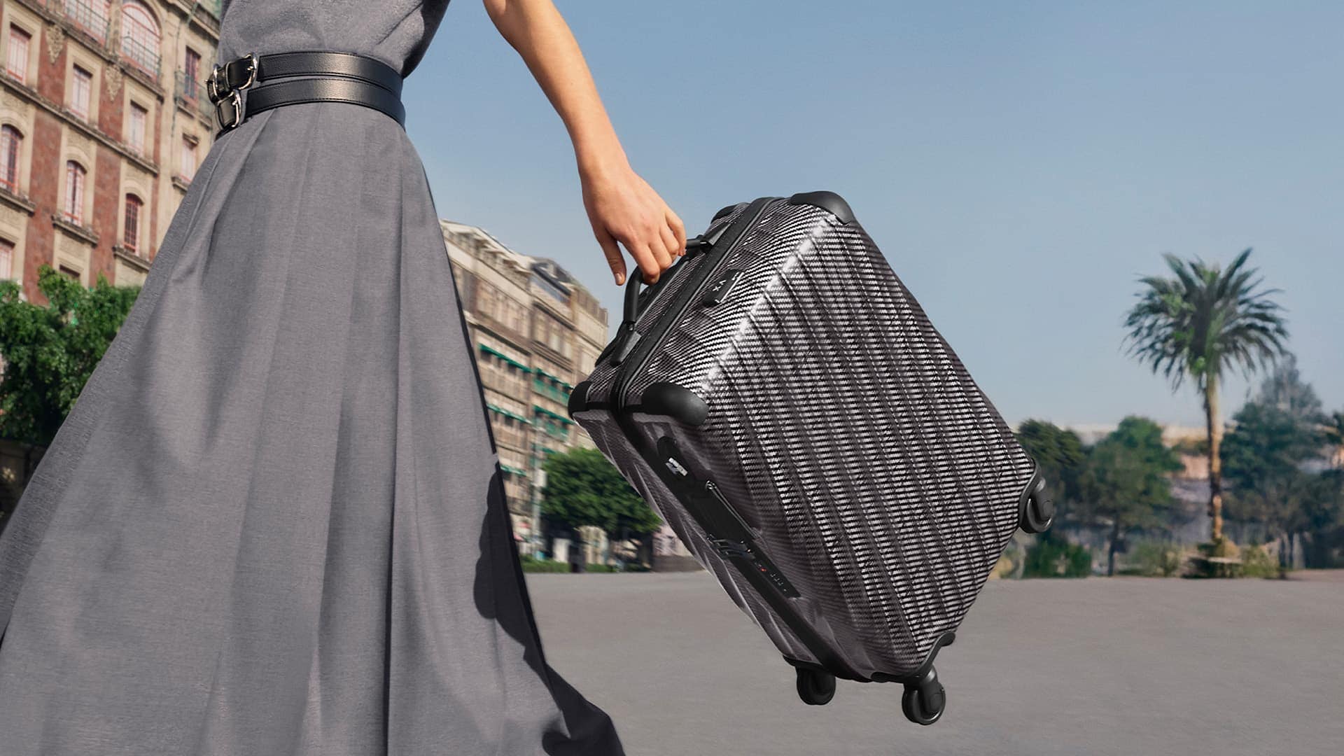 Person in a grey dress carrying a textured dark wheeled suitcase outdoors in a public square