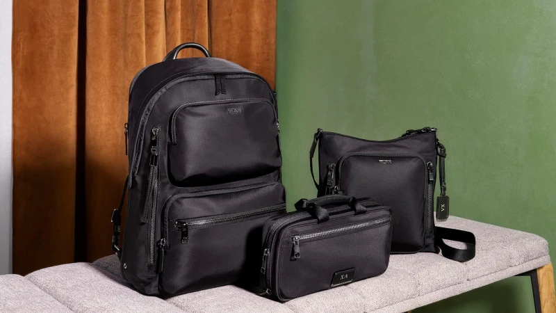 Three black nylon Tumi bags including a backpack, crossbody bag, and pouch displayed on a light gray upholstered bench.