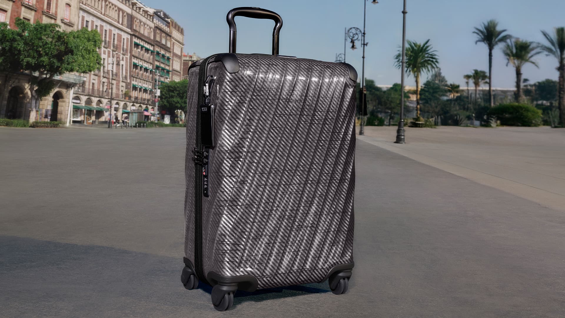 A black carbon fiber hardside suitcase standing upright on a paved plaza with historic buildings and palm trees in the background.