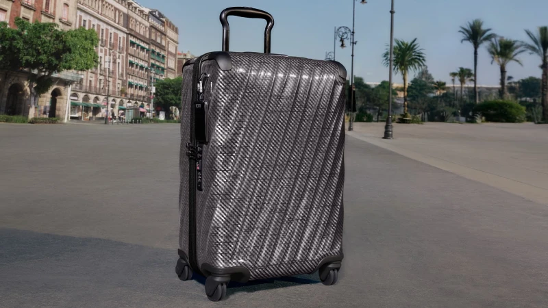A black carbon fiber hardside suitcase standing upright on a paved plaza with historic buildings and palm trees in the background.