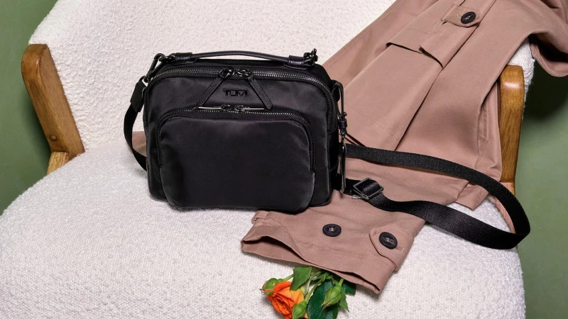 Black nylon Tumi crossbody bag resting on a white textured armchair next to tan garments and an orange rose