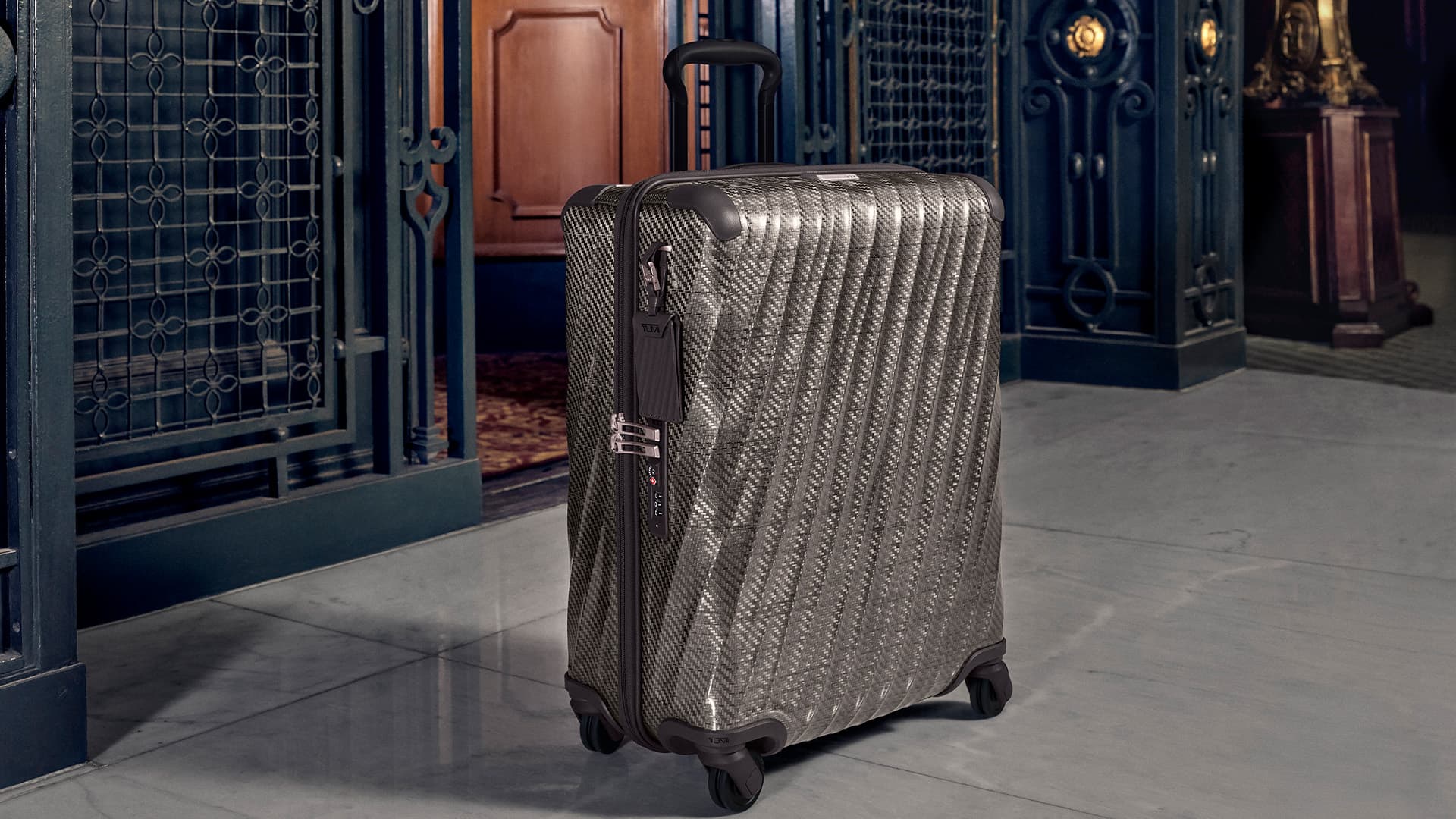 Grey patterned hardside suitcase standing upright on a grey stone floor against an ornate architectural interior.