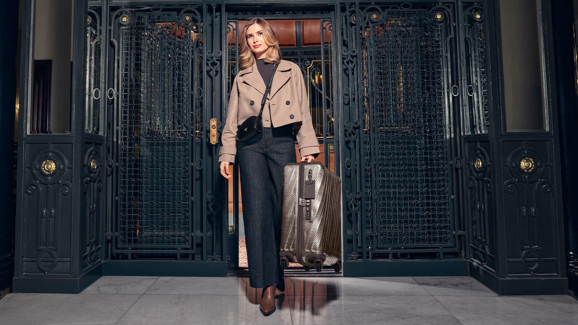 Woman wearing a beige cropped jacket and dark trousers standing before ornate iron doors while pulling a rolling suitcase