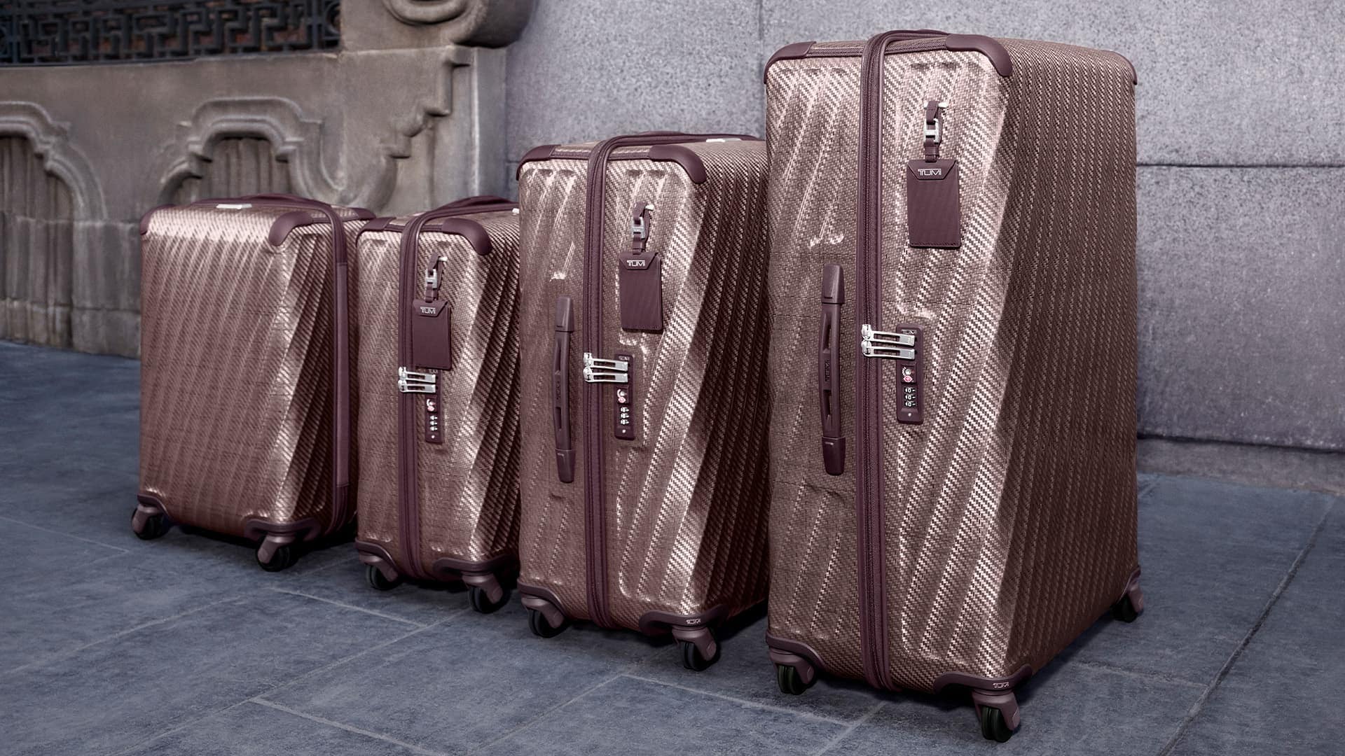 Four matching textured bronze and plum hard-shell suitcases lined up neatly on a stone pavement against a grey wall.