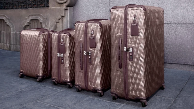 Four matching textured bronze and plum hard-shell suitcases lined up neatly on a stone pavement against a grey wall.