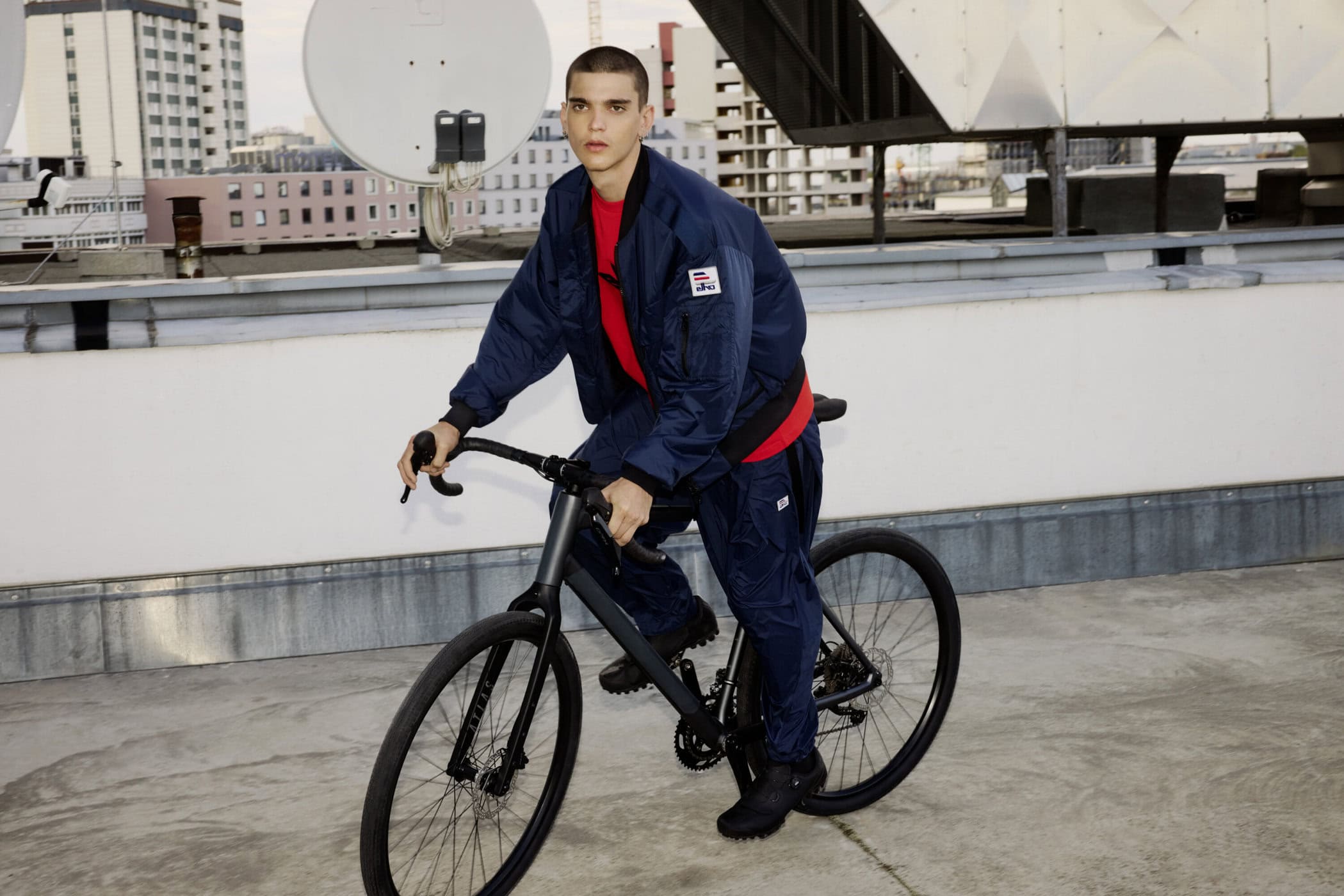 Young man riding a bicycle on a rooftop wearing a navy blue puffer jacket and matching trousers over a red shirt.