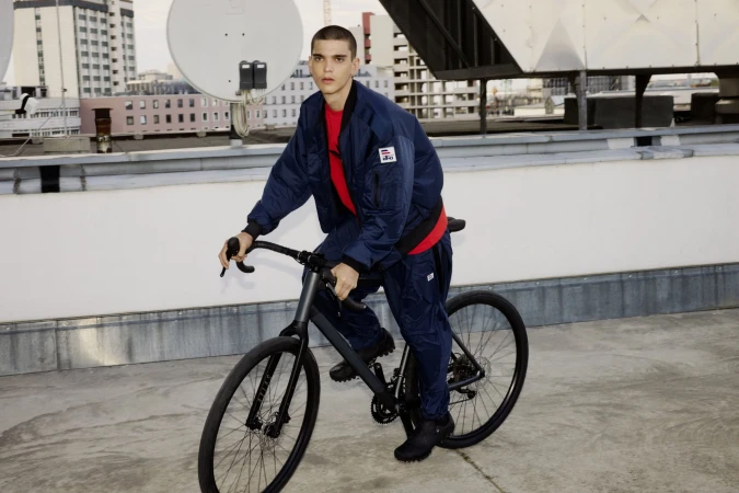 Young man riding a bicycle on a rooftop wearing a navy blue puffer jacket and matching trousers over a red shirt.