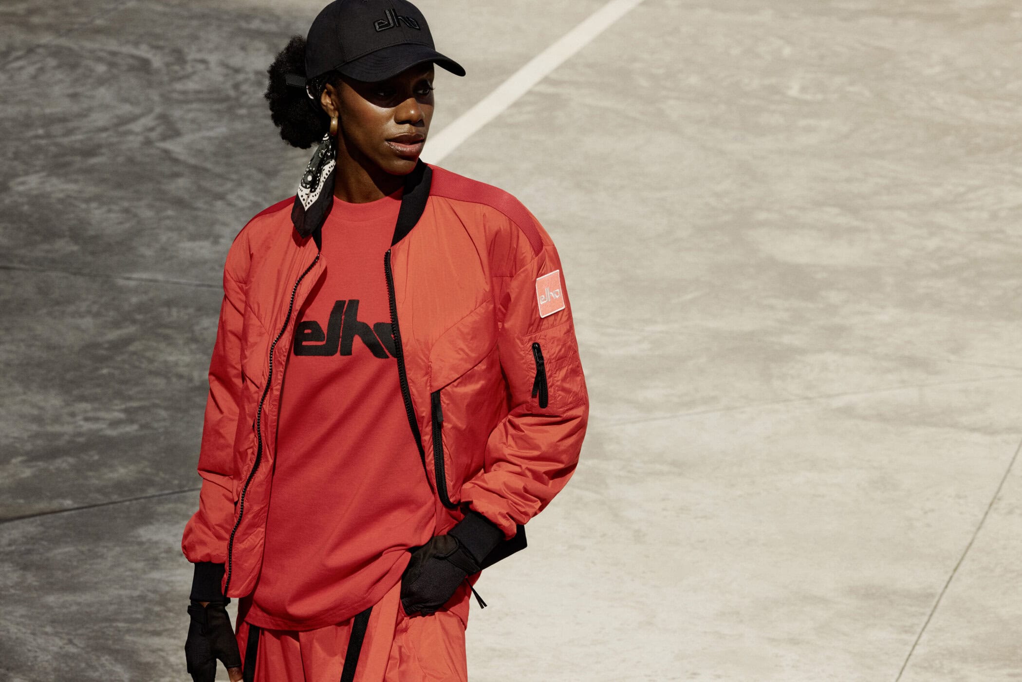 Black woman wearing a red bomber jacket and black cap against a pale court surface