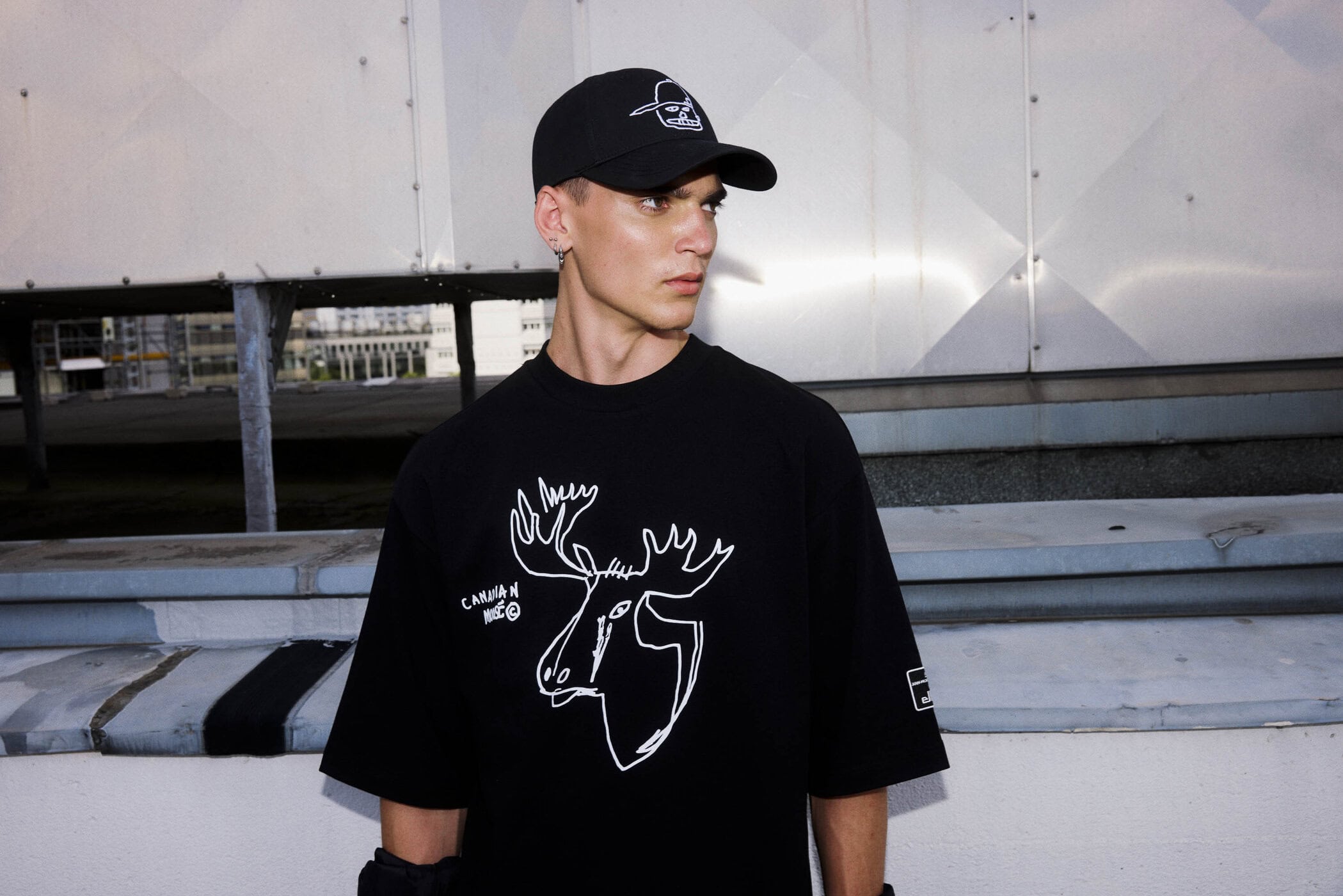 Young man wearing a black cap and graphic t-shirt standing outdoors on a rooftop terrace.