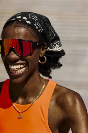 Close-up portrait of a smiling Black woman wearing red mirrored sunglasses, an orange tank top, gold hoop earrings, and a black bandana.