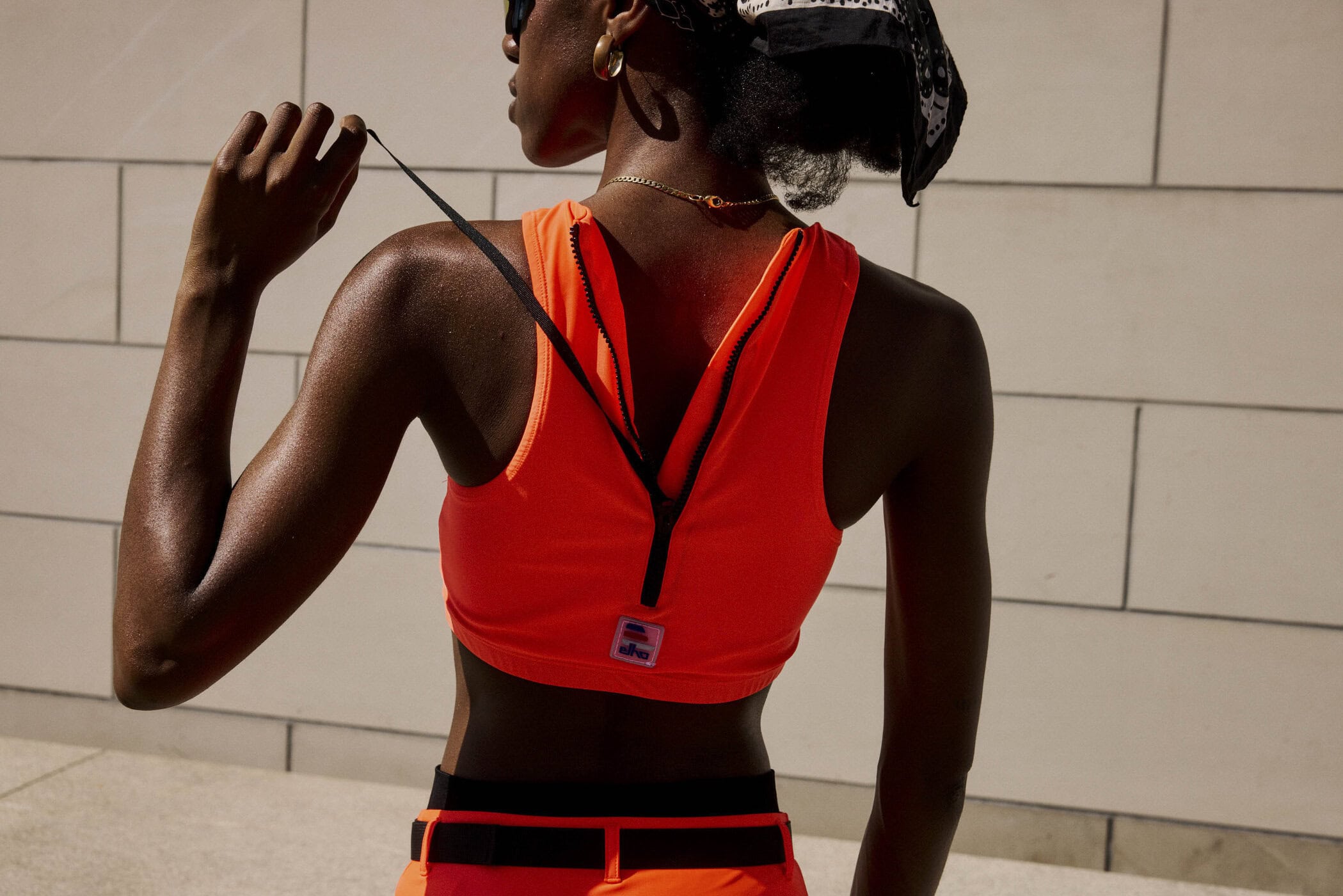 A person wearing a bright orange zip-up top standing against a stone tiled wall for Elho spring 2025 campaign.
