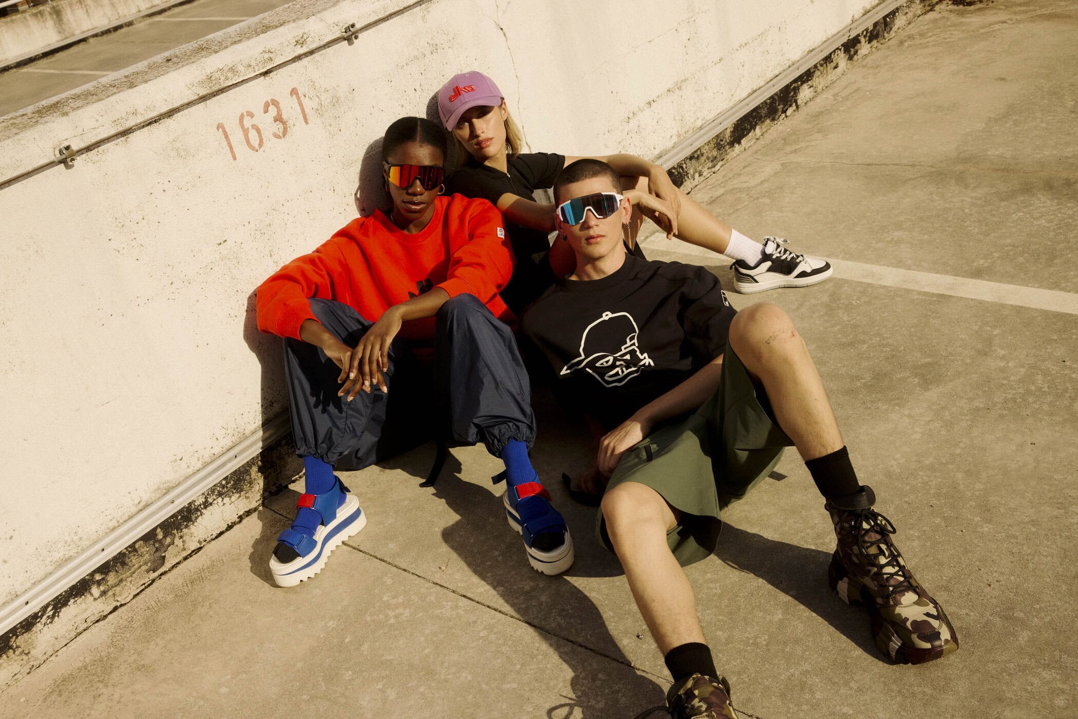 Three models wearing sportswear and sunglasses seated against an outdoor concrete wall for the Elho Spring 2025 campaign.