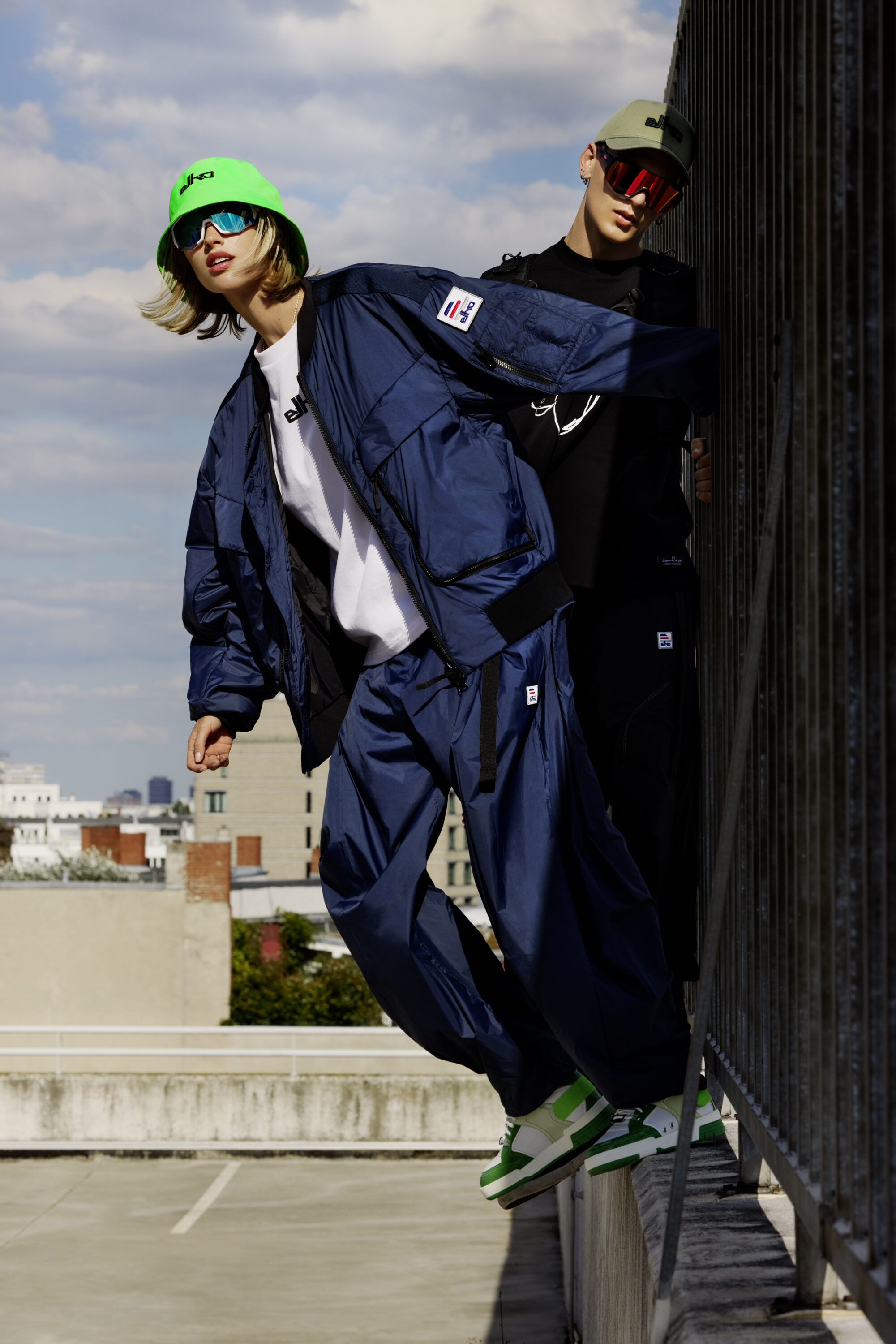 Two models wearing outerwear and bucket hats leaning against a rooftop railing under an open sky.