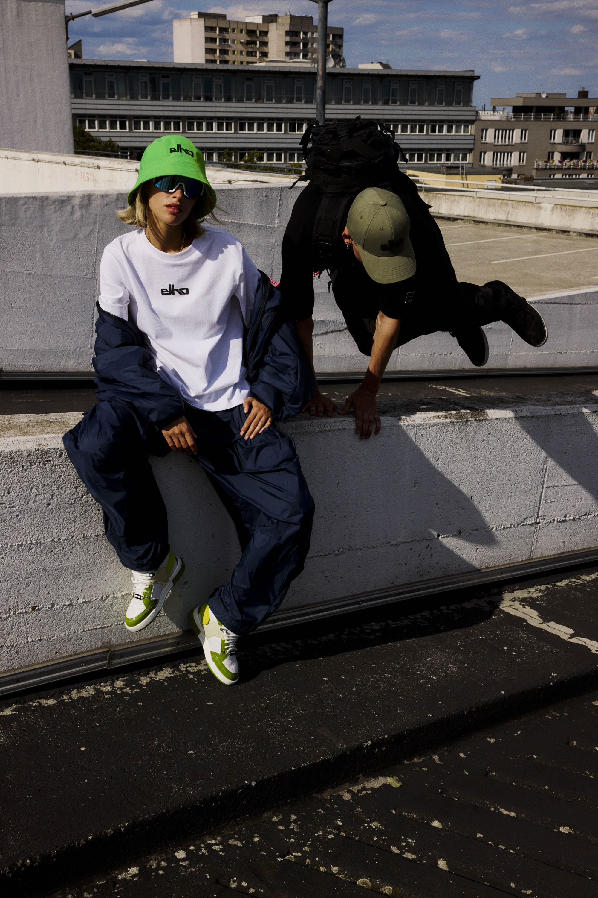 Two models wearing streetwear clothing on a concrete rooftop with urban buildings in the background.