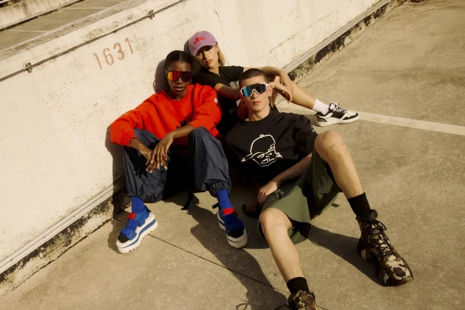 Three models wearing sportswear and sunglasses seated against an outdoor concrete wall for the Elho Spring 2025 campaign.