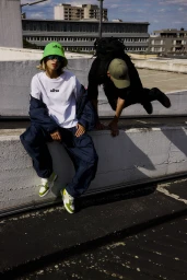 Two models wearing streetwear clothing on a concrete rooftop with urban buildings in the background.