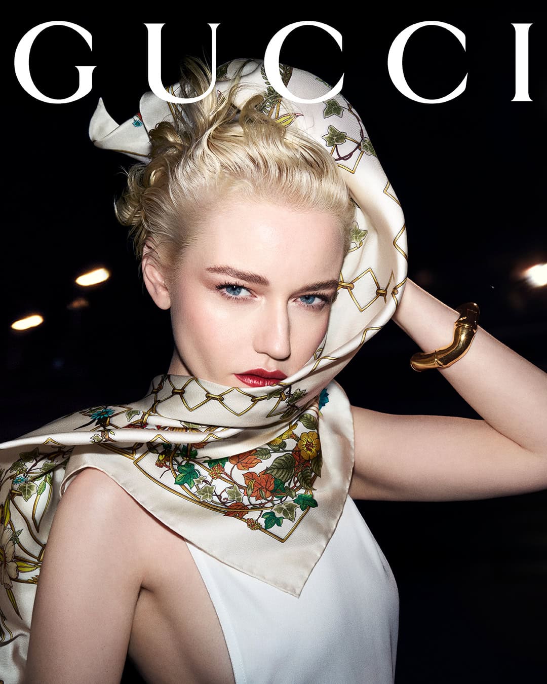 Blonde woman wearing a patterned silk scarf over her head and a white top with bold GUCCI text above.