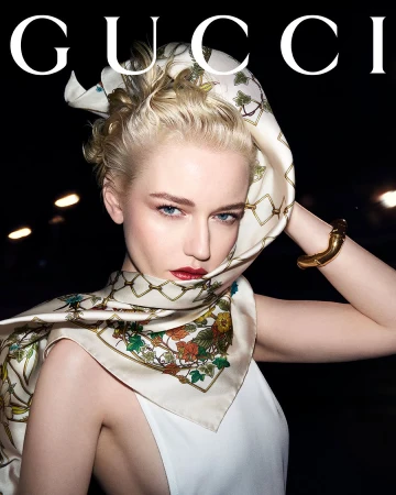 Blonde woman wearing a patterned silk scarf over her head and a white top with bold GUCCI text above.