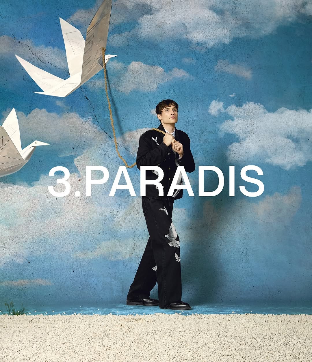 Model in black jacket and trousers holding a rope attached to large origami paper birds against a painted cloud backdrop.
