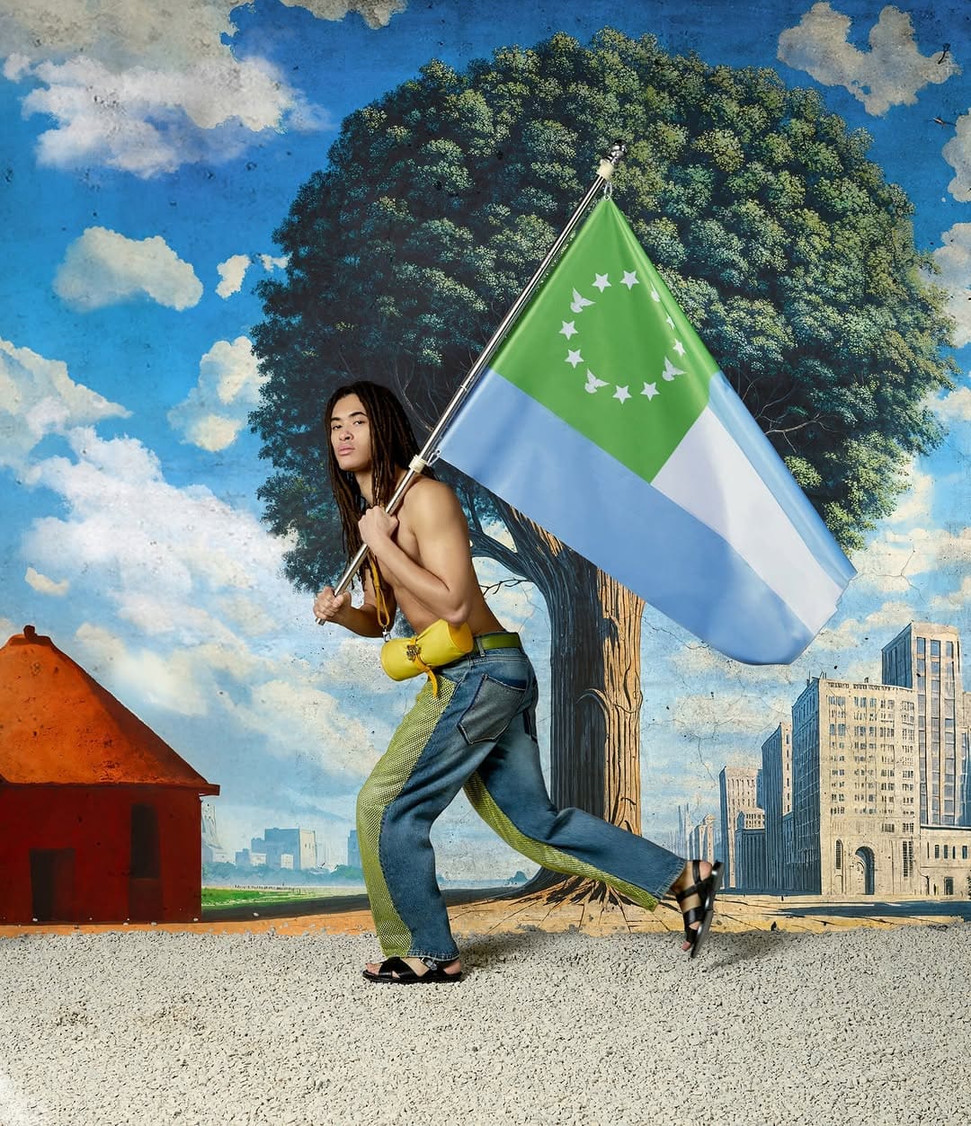 shirtless male model with dreadlocks carrying a large green and blue flag against a painted background of a tree and buildings.