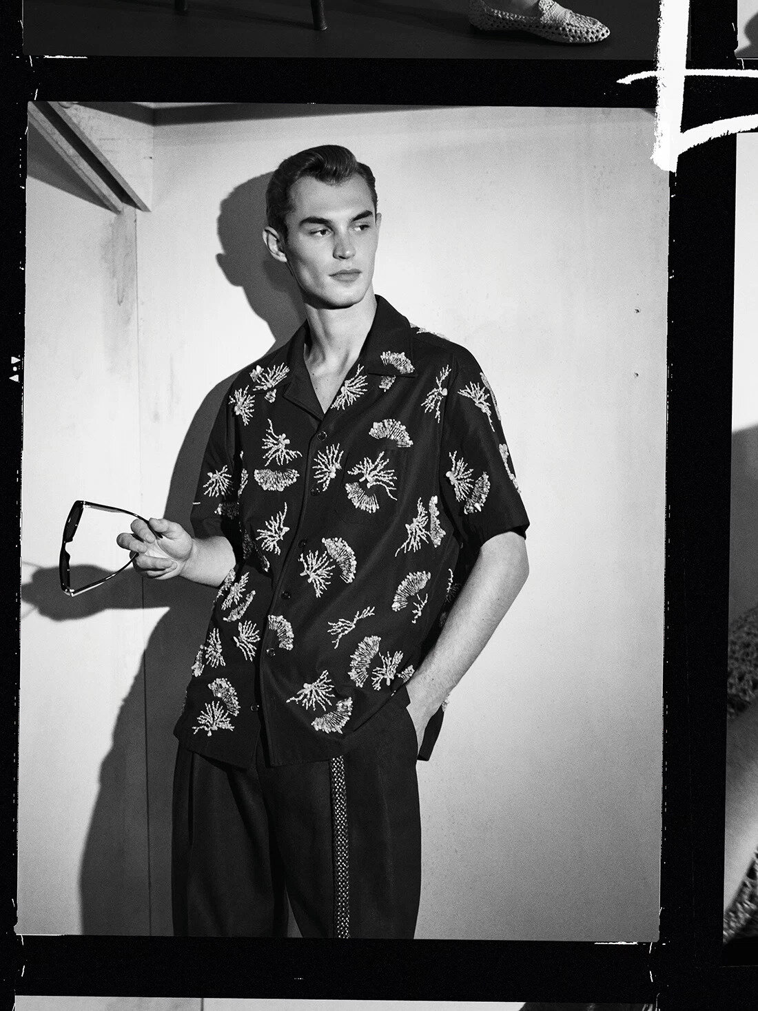 Model Kit Butler wearing a patterned short-sleeve shirt holding sunglasses in a black-and-white fashion campaign photograph.