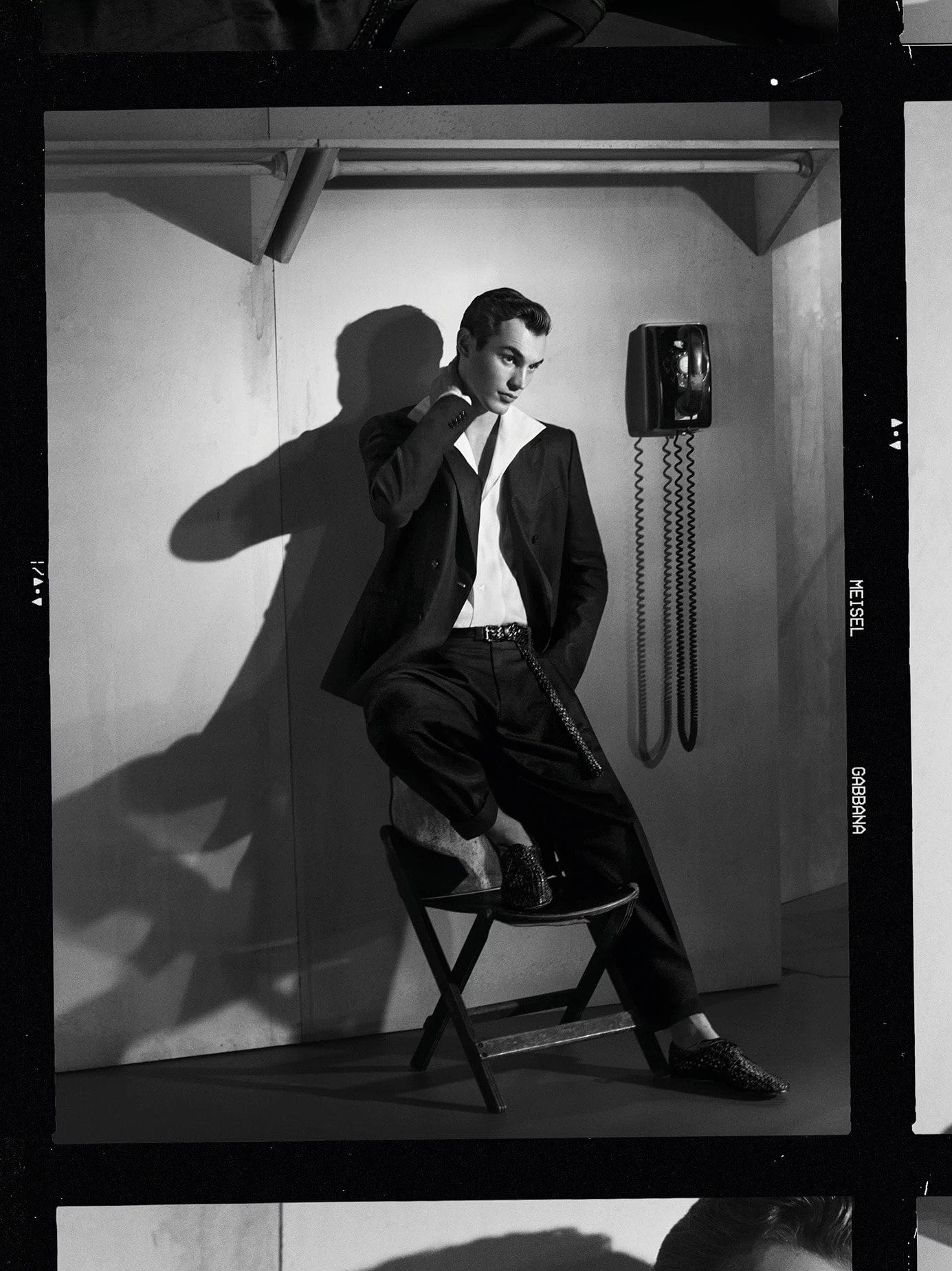 Model Kit Butler in a dark suit sitting on a chair next to a wall phone for Dolce and Gabbana Spring 2025 campaign.