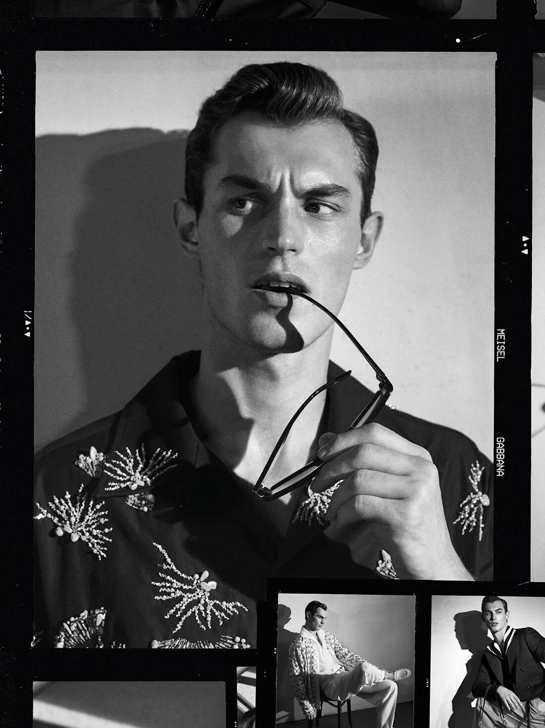 Black and white portrait of a male model holding sunglasses to his mouth, wearing an embroidered shirt for Dolce & Gabbana.