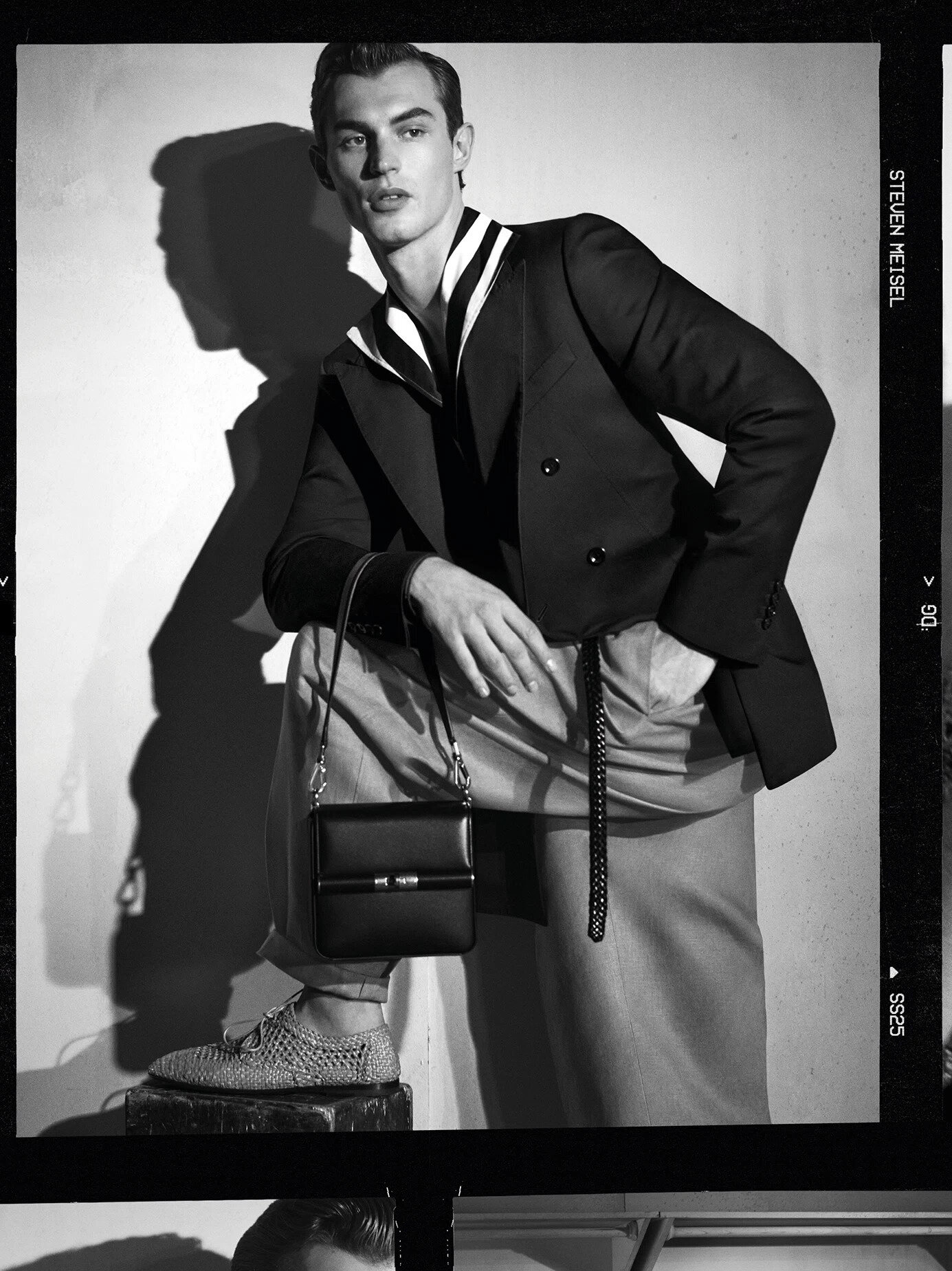 Black and white portrait of a male model posing in a tailored jacket and holding a handbag against a plain background