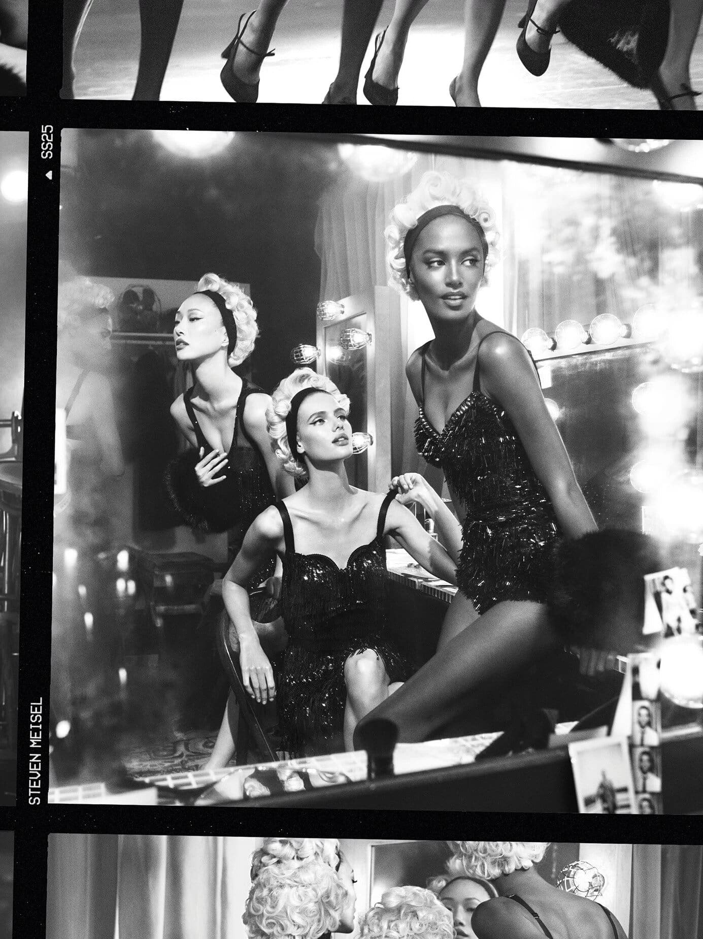 Black and white editorial image of multiple female models wearing sparkling bodysuits in a backstage dressing room setting.