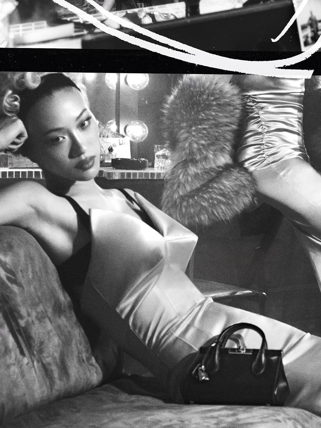 Model lounging on a sofa wearing a structured white dress with a black handbag nearby in a monochrome setting