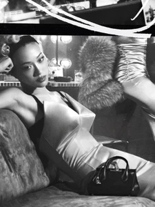 Model lounging on a sofa wearing a structured white dress with a black handbag nearby in a monochrome setting