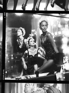 Black and white editorial image of multiple female models wearing sparkling bodysuits in a backstage dressing room setting.