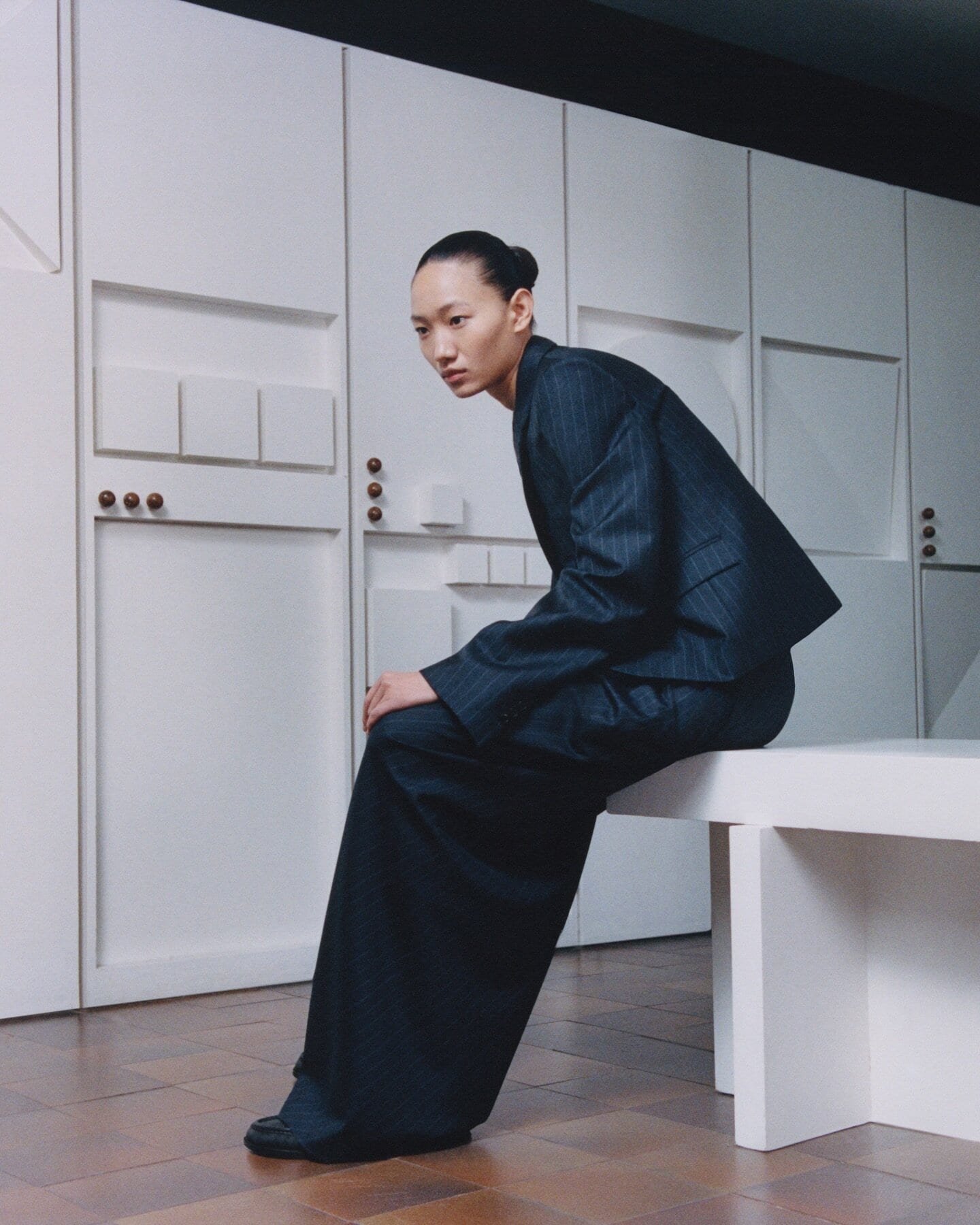 A model seated on a white bench wearing a dark structured suit against a minimalist white wall with paneling.