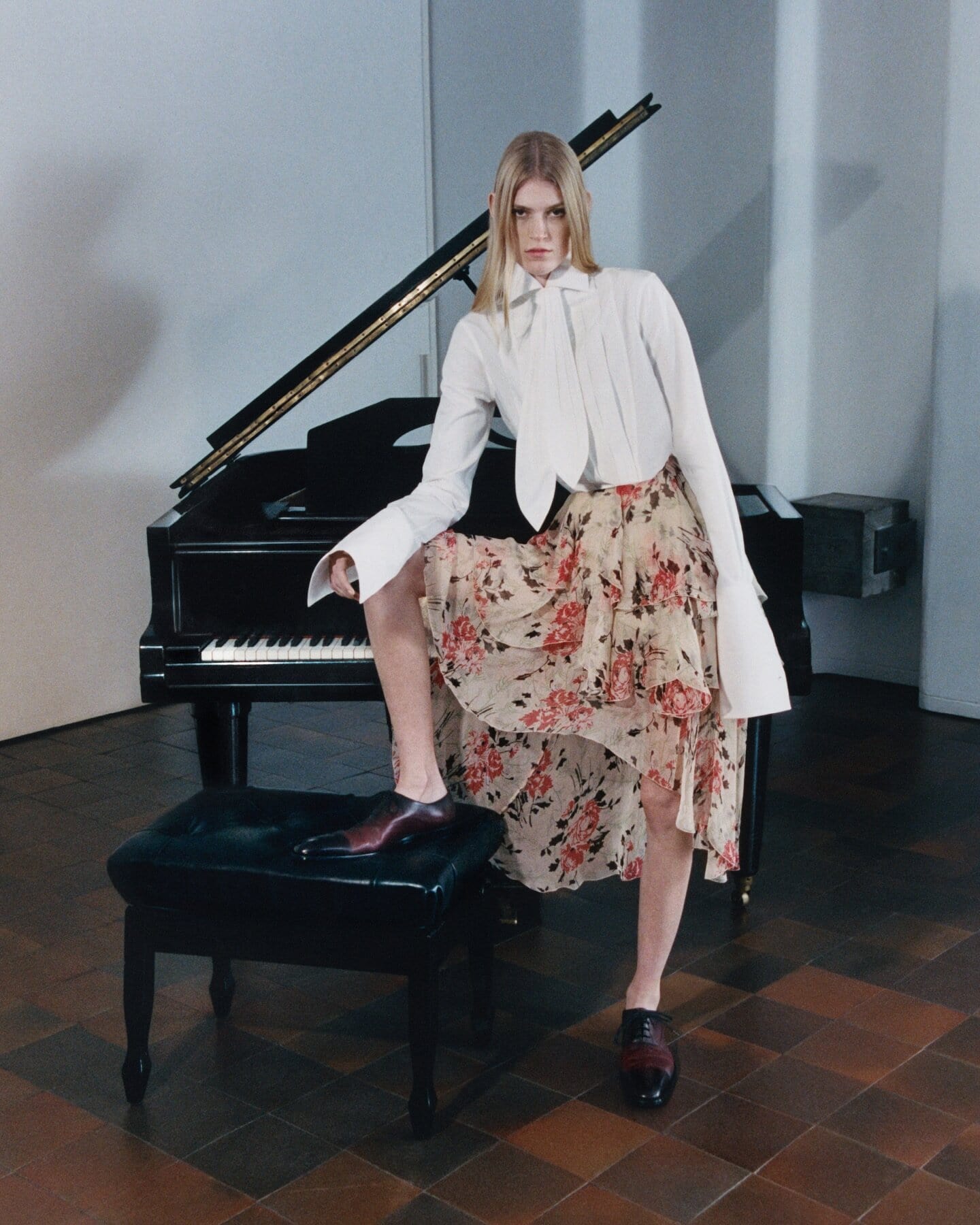 Model wearing white blouse and floral skirt posing with one foot on a piano bench next to a grand piano