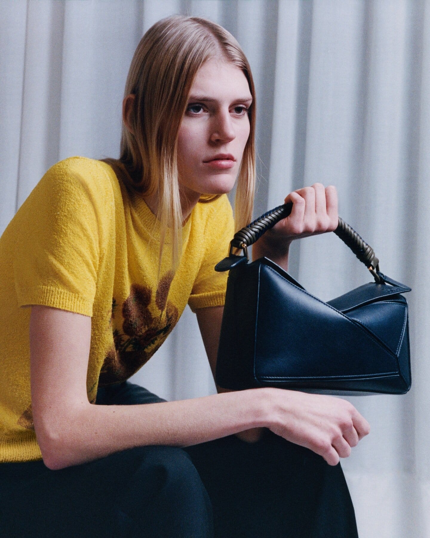 Model in a yellow patterned top holding a dark leather handbag against white draped curtains