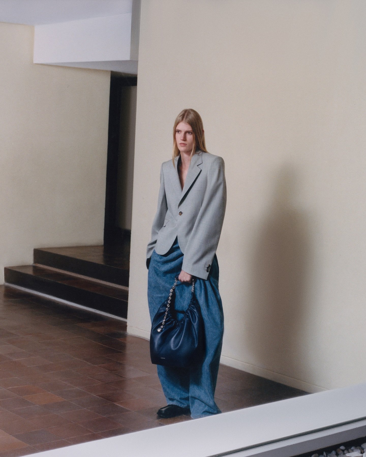 Model standing in an interior space wearing a grey blazer, wide blue trousers, and holding a dark handbag.