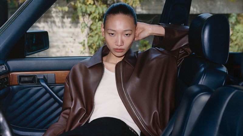 Model seated inside a car wearing a brown leather jacket and white top, looking directly at the camera.