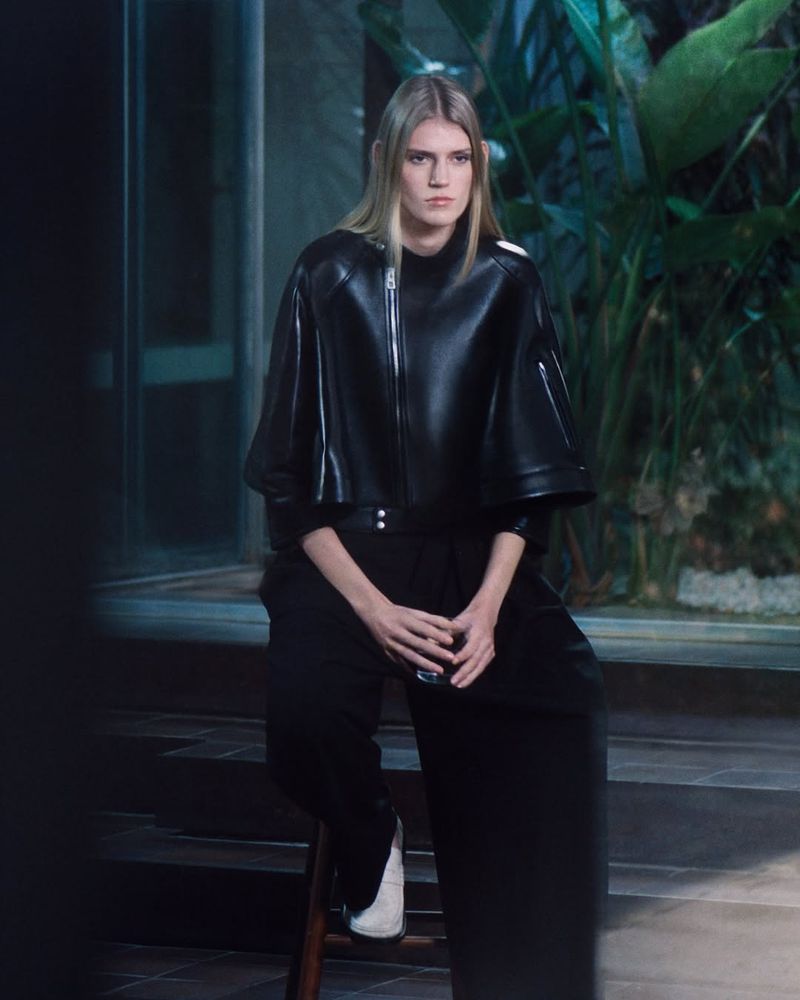 Model with long blonde hair seated outdoors wearing a black leather jacket and black trousers