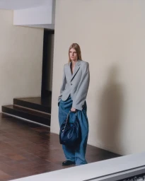 Model standing in an interior space wearing a grey blazer, wide blue trousers, and holding a dark handbag.