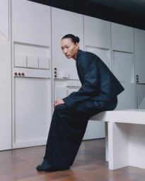 A model seated on a white bench wearing a dark structured suit against a minimalist white wall with paneling.