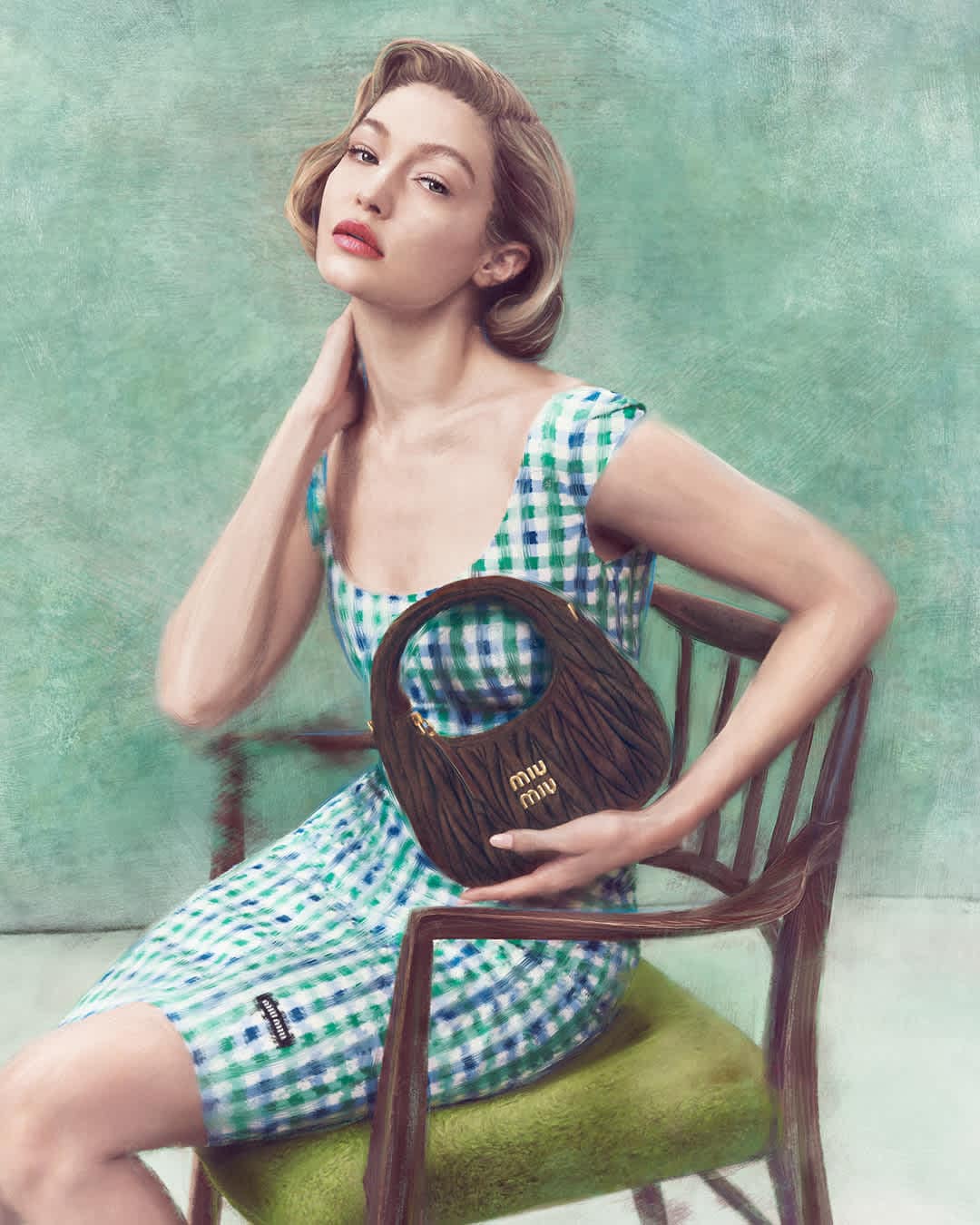 Gigi Hadid seated in a wooden chair holding a dark gathered leather handbag for Miu Miu
