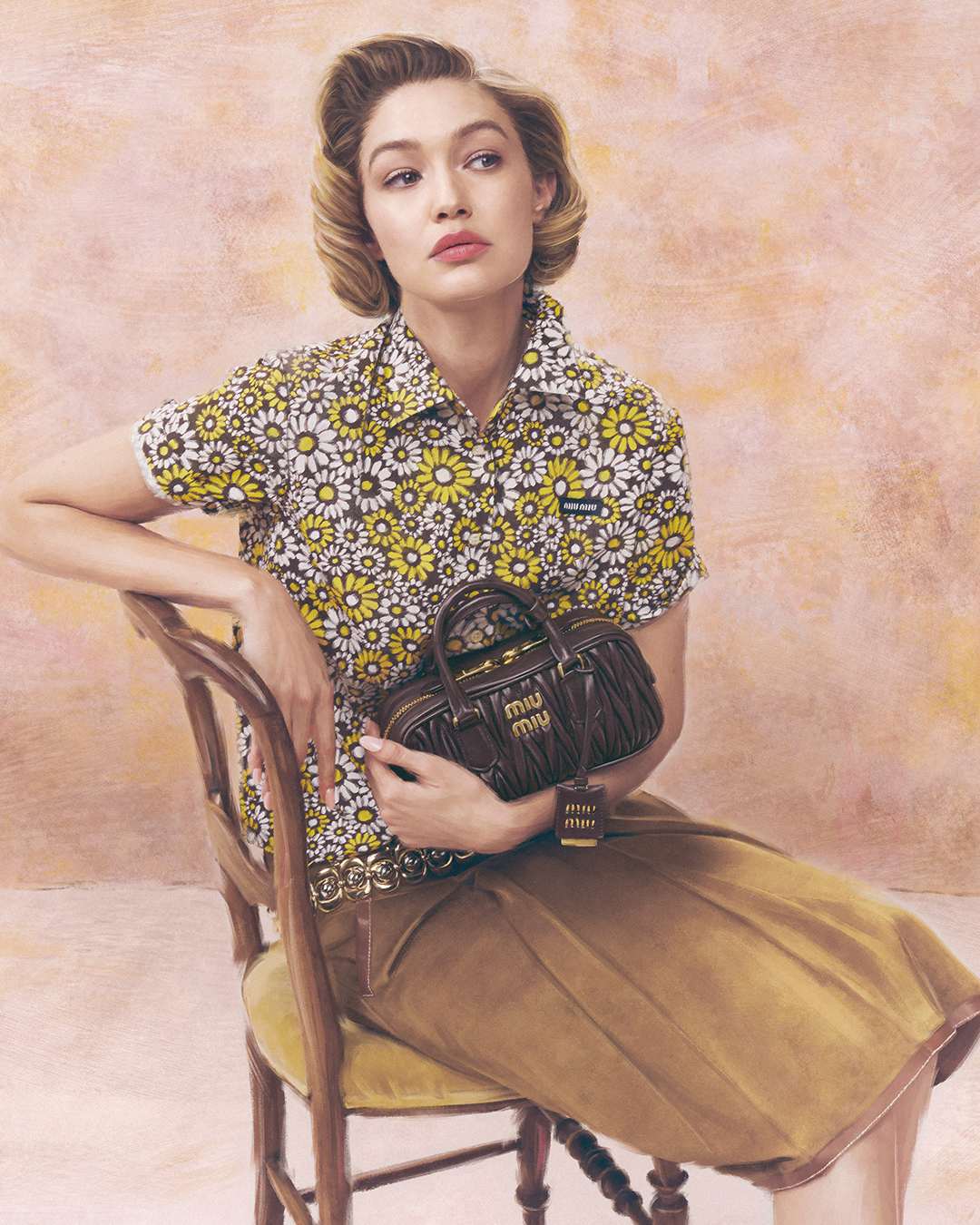 Gigi Hadid wearing a floral patterned short-sleeve shirt and holding a brown leather handbag while seated on a wooden chair.