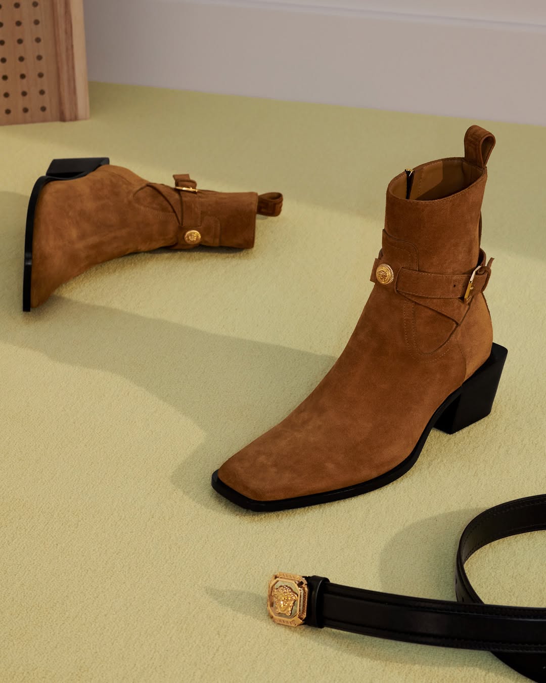 Tan suede ankle boots with buckle straps and black block heels resting on a pale green surface.