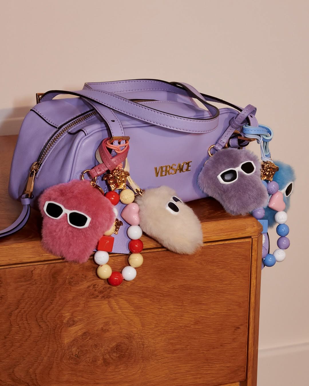 Lavender Versace shoulder bag adorned with fuzzy charms and colorful bead straps resting on a wooden surface.