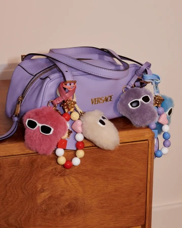 Lavender Versace shoulder bag adorned with fuzzy charms and colorful bead straps resting on a wooden surface.